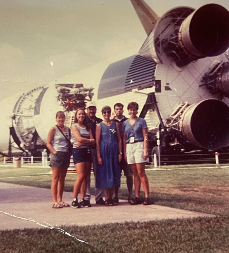A family of 6 stands poses together in front of a rocket
