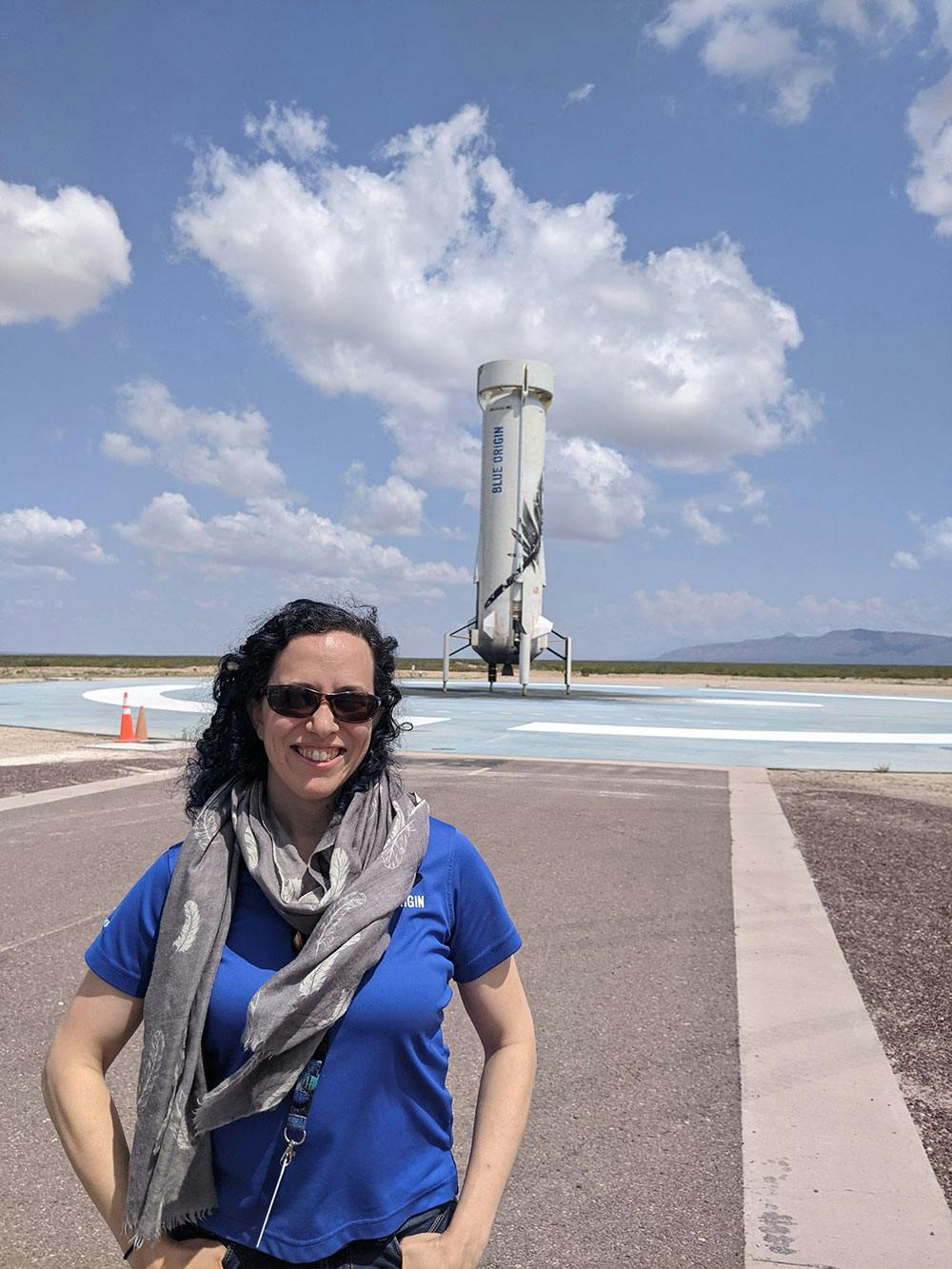 A woman in a blue shirt and gray scarf stands in front of the New Shepard booster on the landing pad