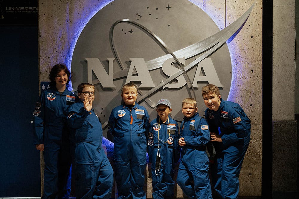 A group of kids in blue NASA jumpsuits pose by a NASA logo sign