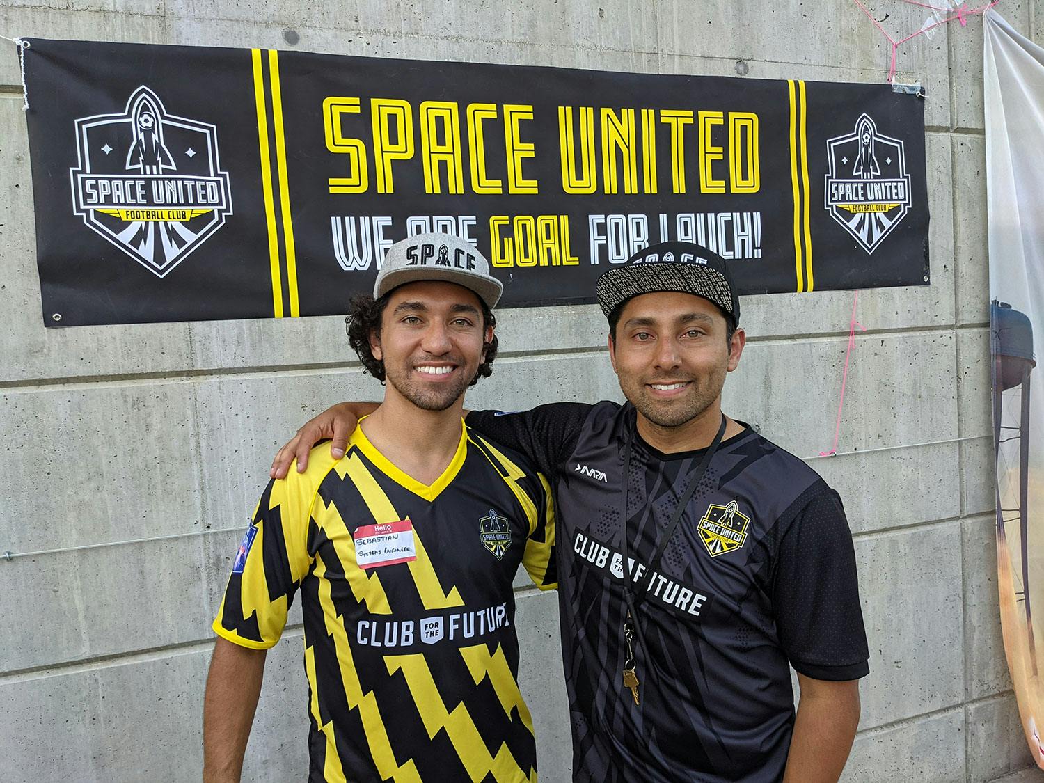 Two men in soccer jerseys and hats in front of a 'Space United' sign